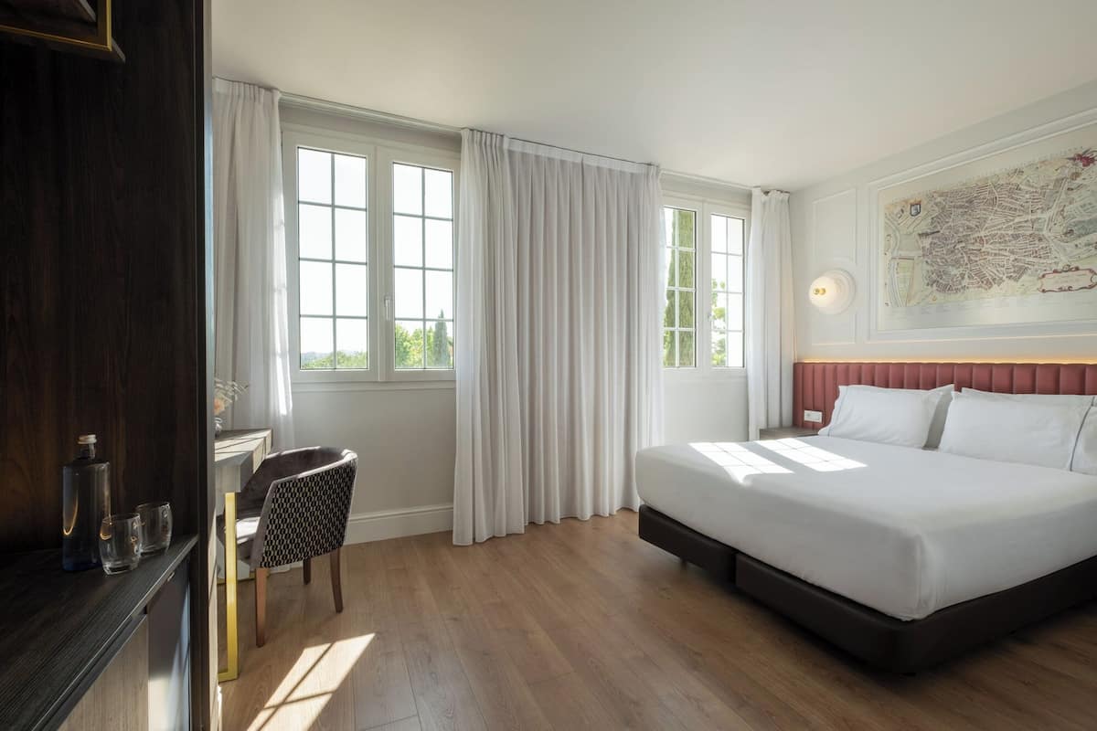 Une chambre d’hôtel avec un grand lit, deux fenêtres avec des rideaux, une carte accrochée au mur et une petite table avec des verres.