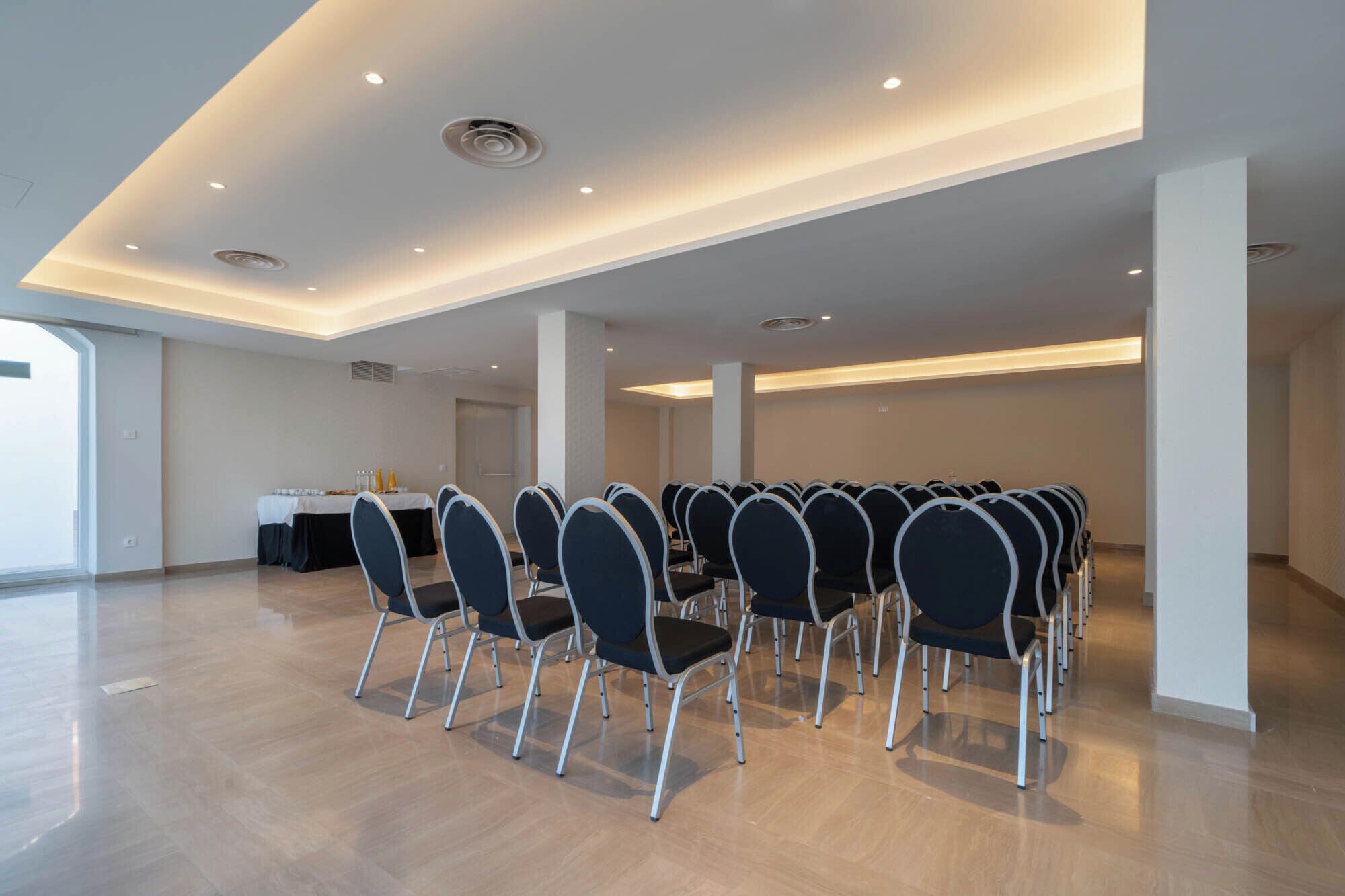 Un salón de conferencias con filas de sillas, una mesa cubierta con mantel blanco y una puerta al fondo.
