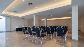 Un salón de conferencias con filas de sillas, una mesa cubierta con mantel blanco y una puerta al fondo.