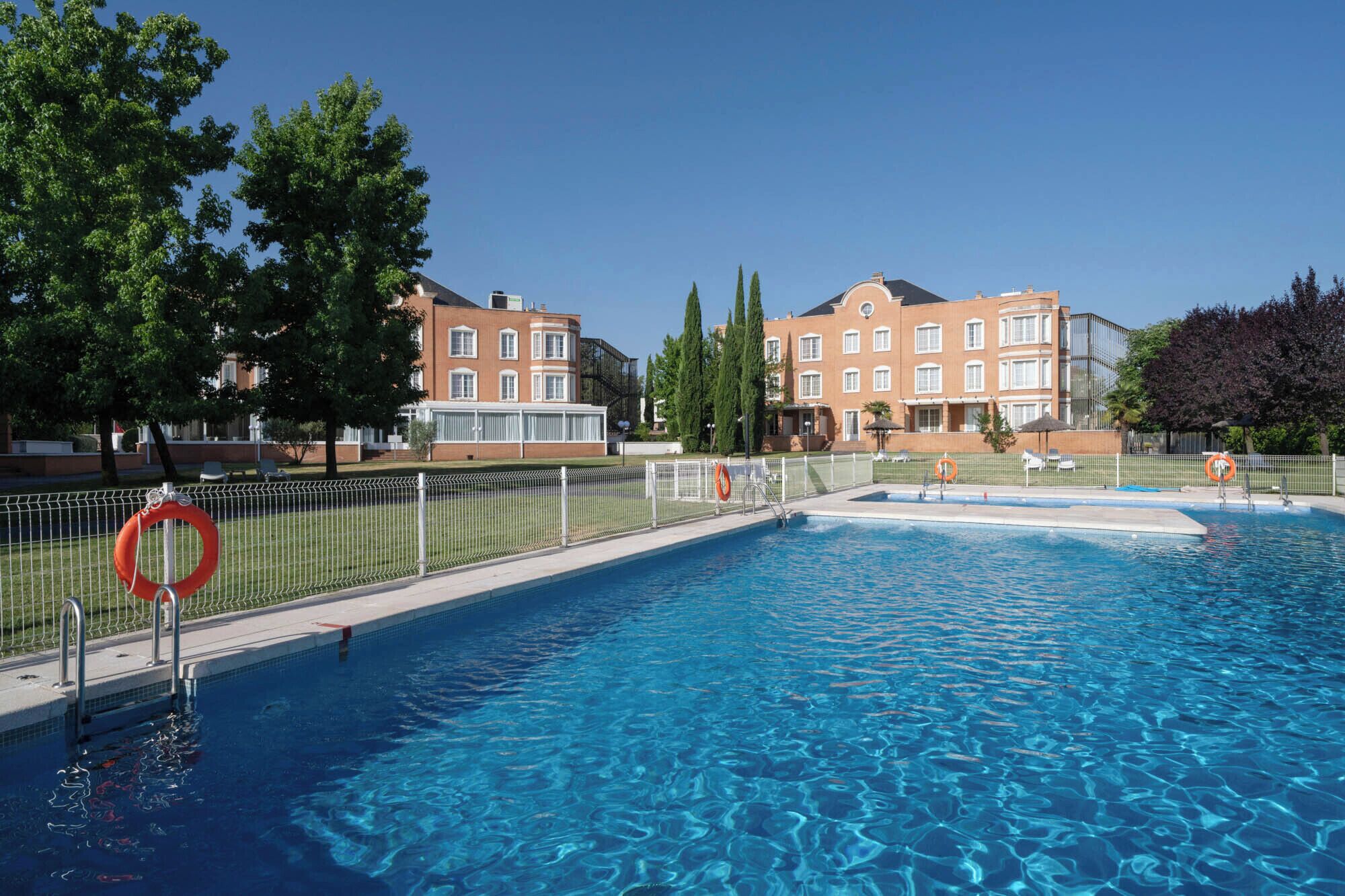 Uma piscina com um salva-vidas, cercada por uma grade, e edifícios ao fundo.