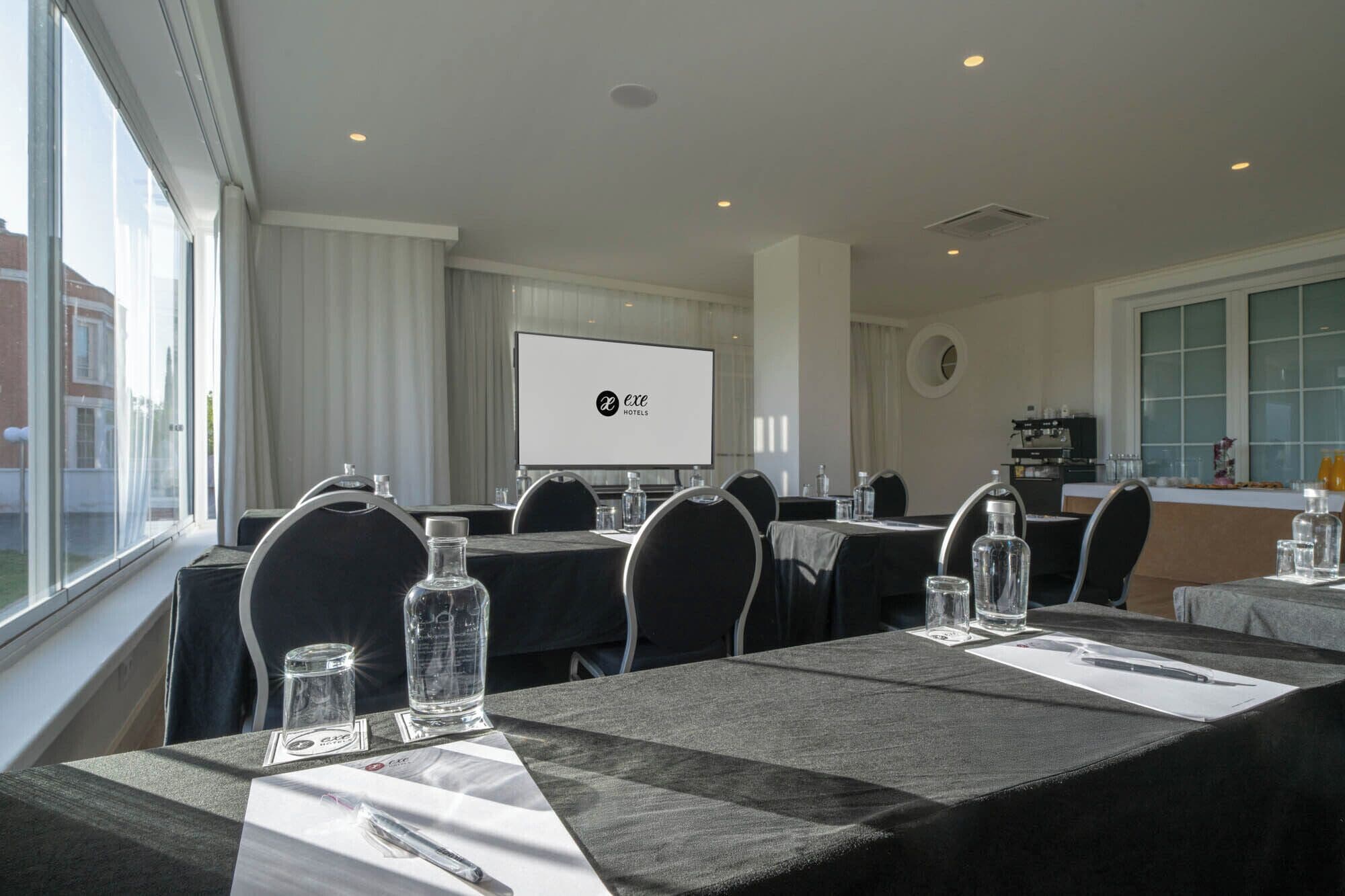 A conference room with tables, chairs, a projector screen, and a kitchen area.