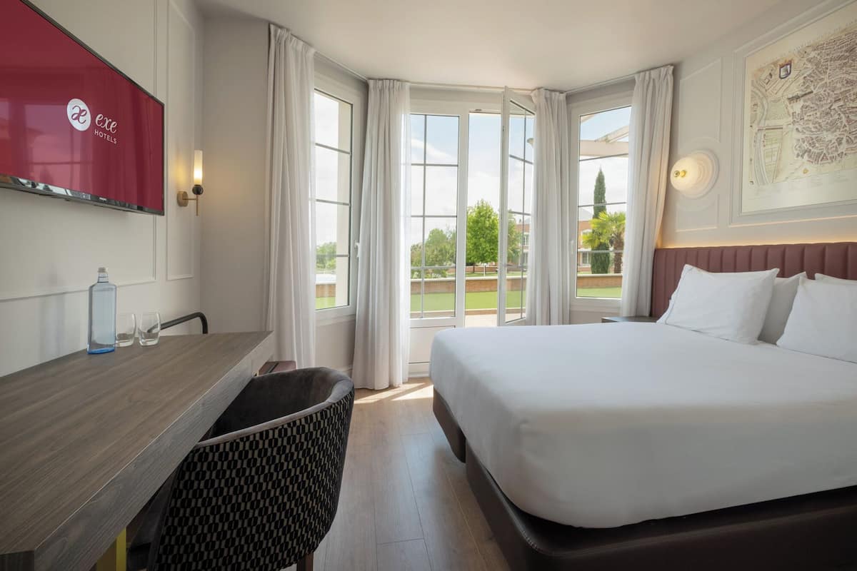 Une chambre d’hôtel équipée d’un lit, d’un bureau, d’une chaise, d’une télévision et de grandes fenêtres.