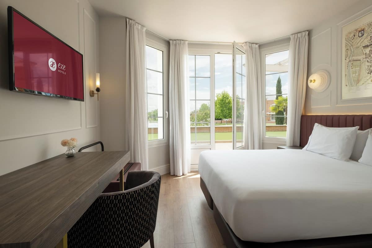 Habitación de hotel con cama, escritorio, silla, televisor y ventanas amplias.