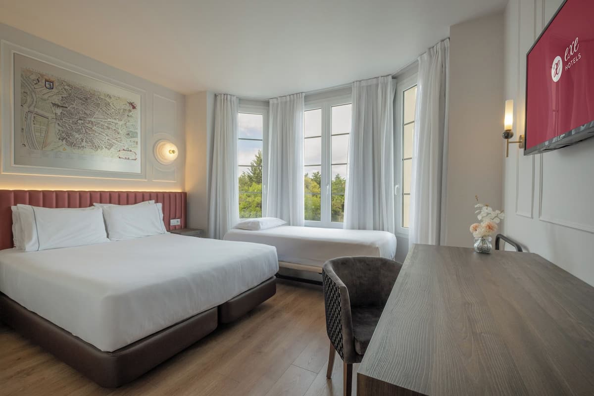 Une chambre d’hôtel avec deux lits, une table à manger, une chaise, une carte accrochée au mur et une vue sur la verdure à l’extérieur.