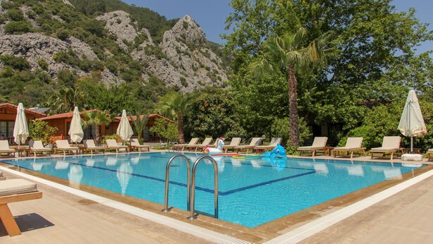 A swimming pool with lounge chairs and umbrellas, surrounded by trees and mountains. - Dolunay Apart Otel (Kemer)
