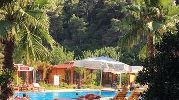 Zona de piscina con sillones de descanso y sombrillas, rodeada de exuberante vegetación y un edificio de madera.