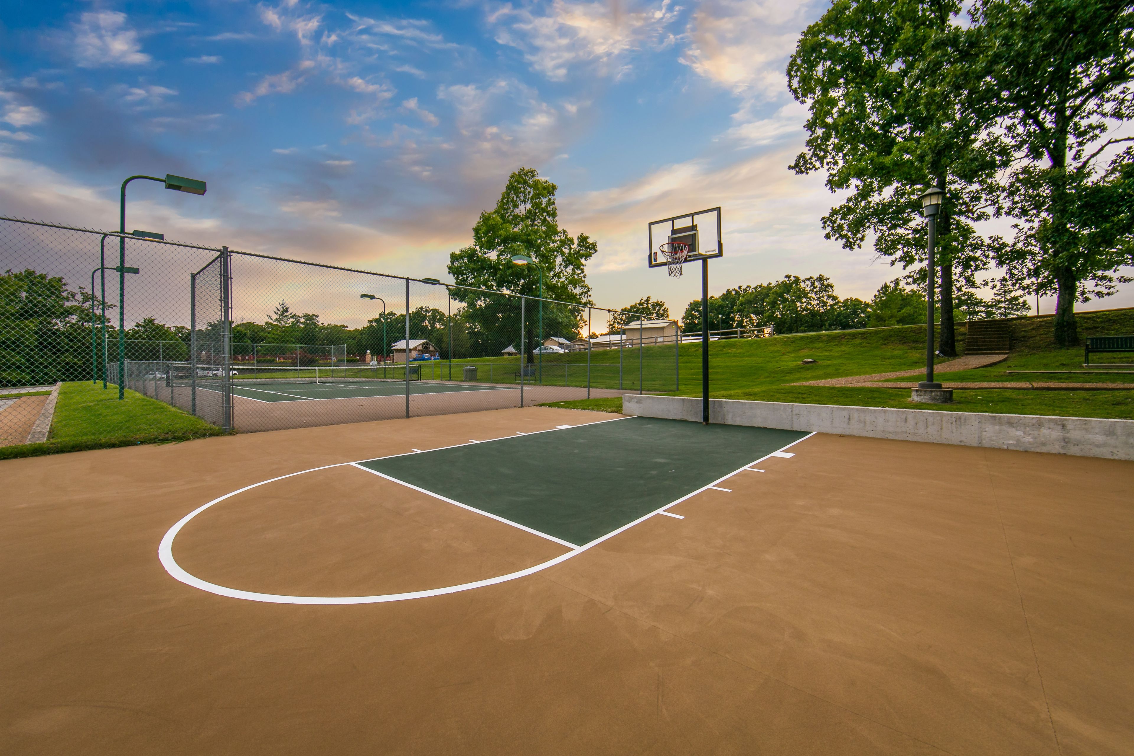 An outdoor basketball court with a green playing surface, surrounded by a chain-link fence, trees, and a clear sky.