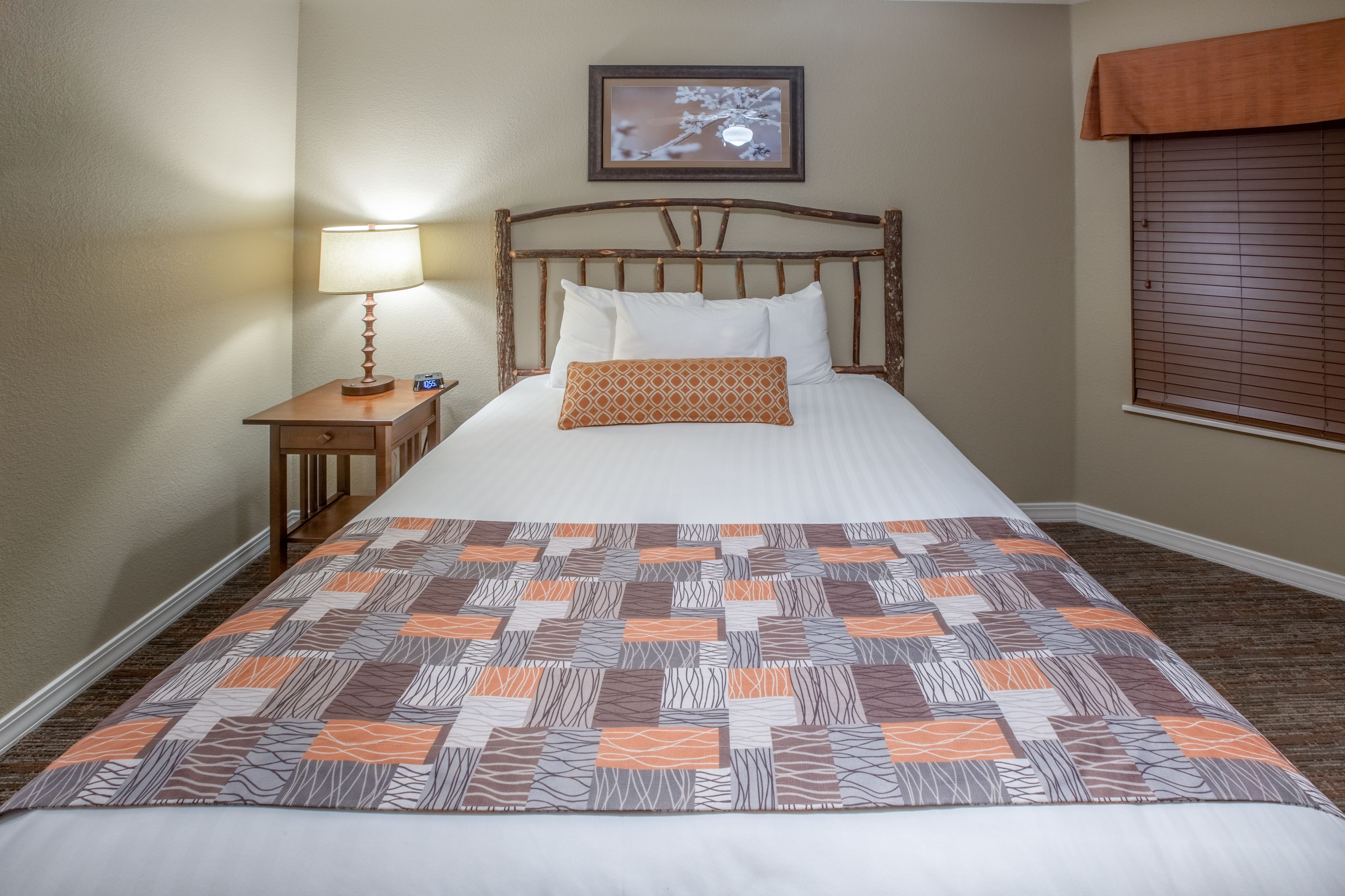 Un letto rifatto con cura, una coperta a motivi decorativi, una lampada da comodino, un quadro appeso alla parete e una finestra con persiane.
