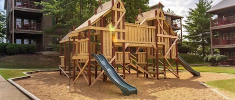 A wooden play structure with a slide, surrounded by a playground area with mulch, set against a backdrop of residential buildings.