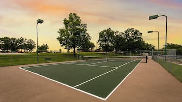 An outdoor tennis court with a net, surrounded by a fence and trees.