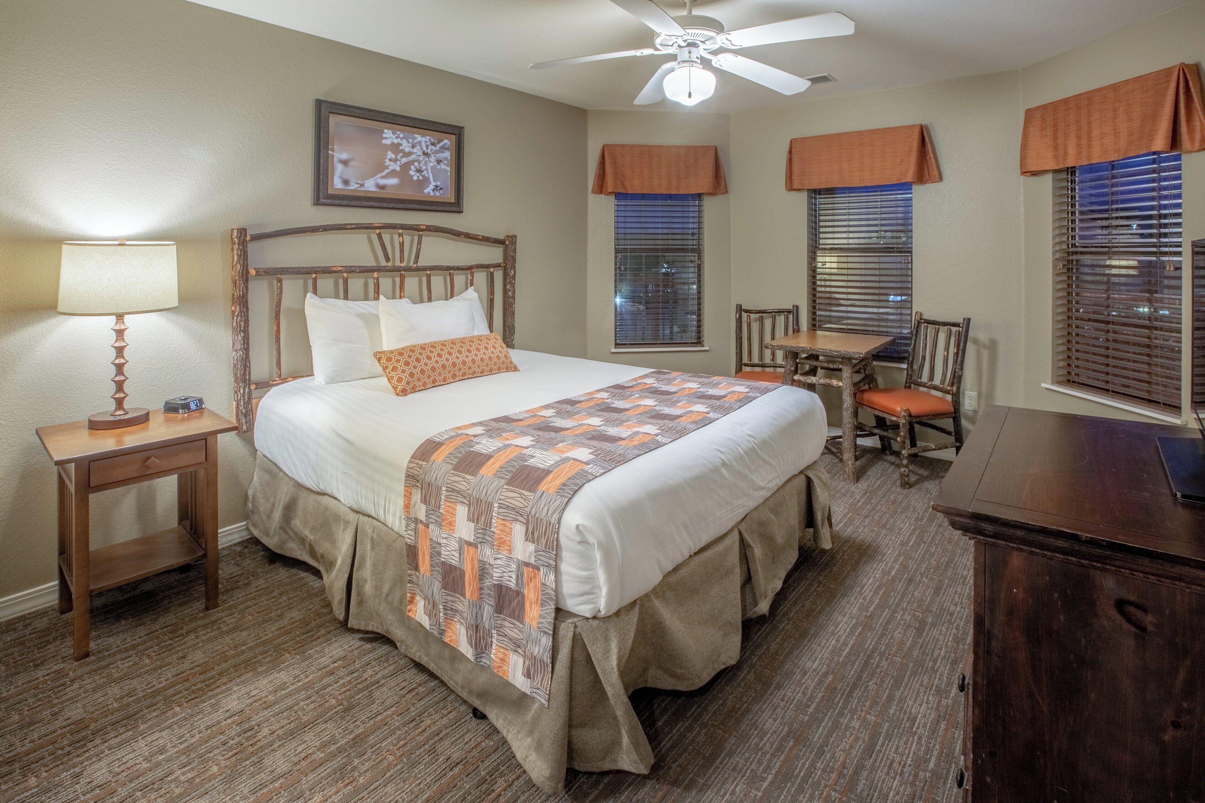 A hotel room with a bed, a desk, a chair, a lamp, a ceiling fan, and a window with blinds.