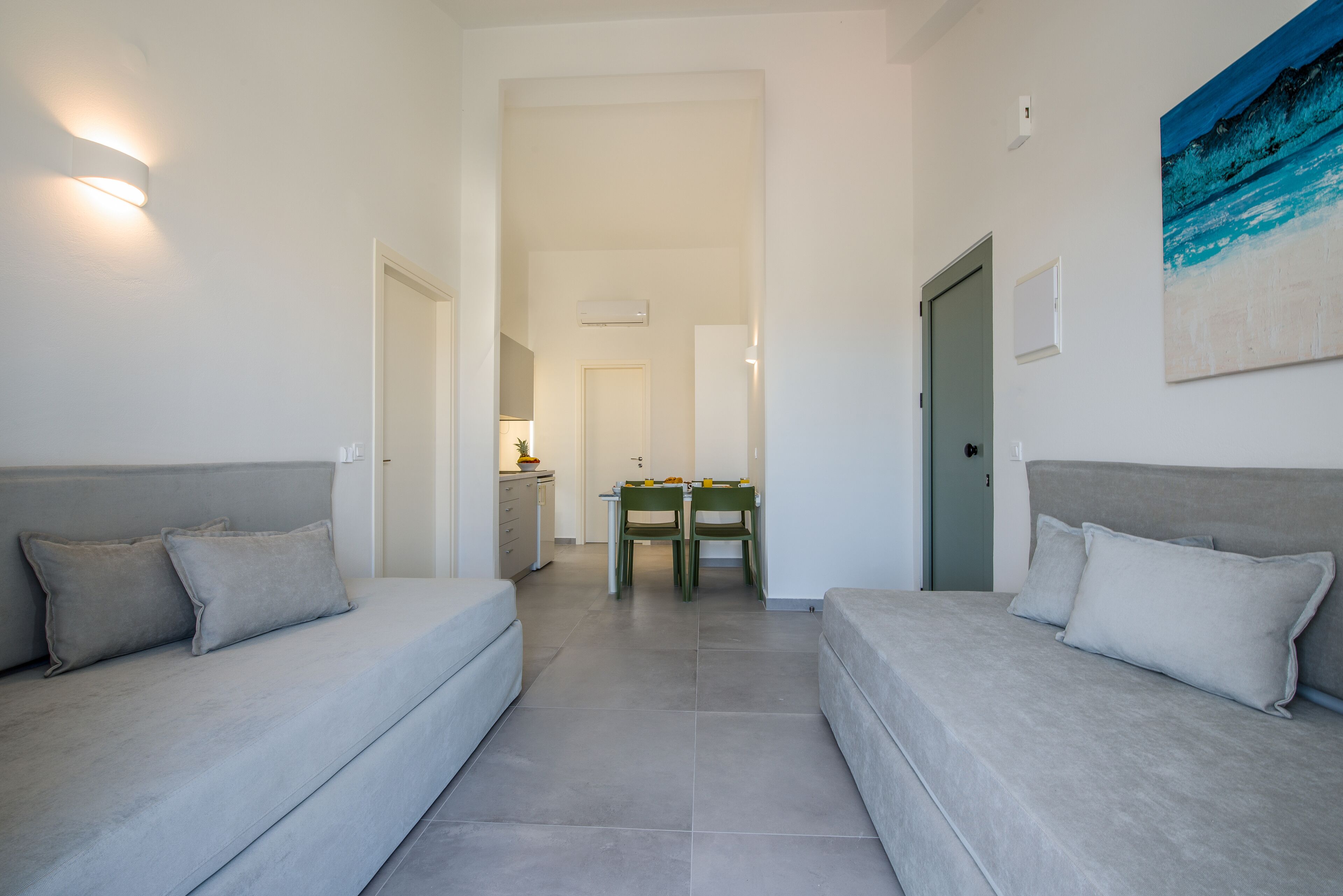 A modern, minimalist room with two grey sofas, a dining table with chairs, and a door leading to another room.