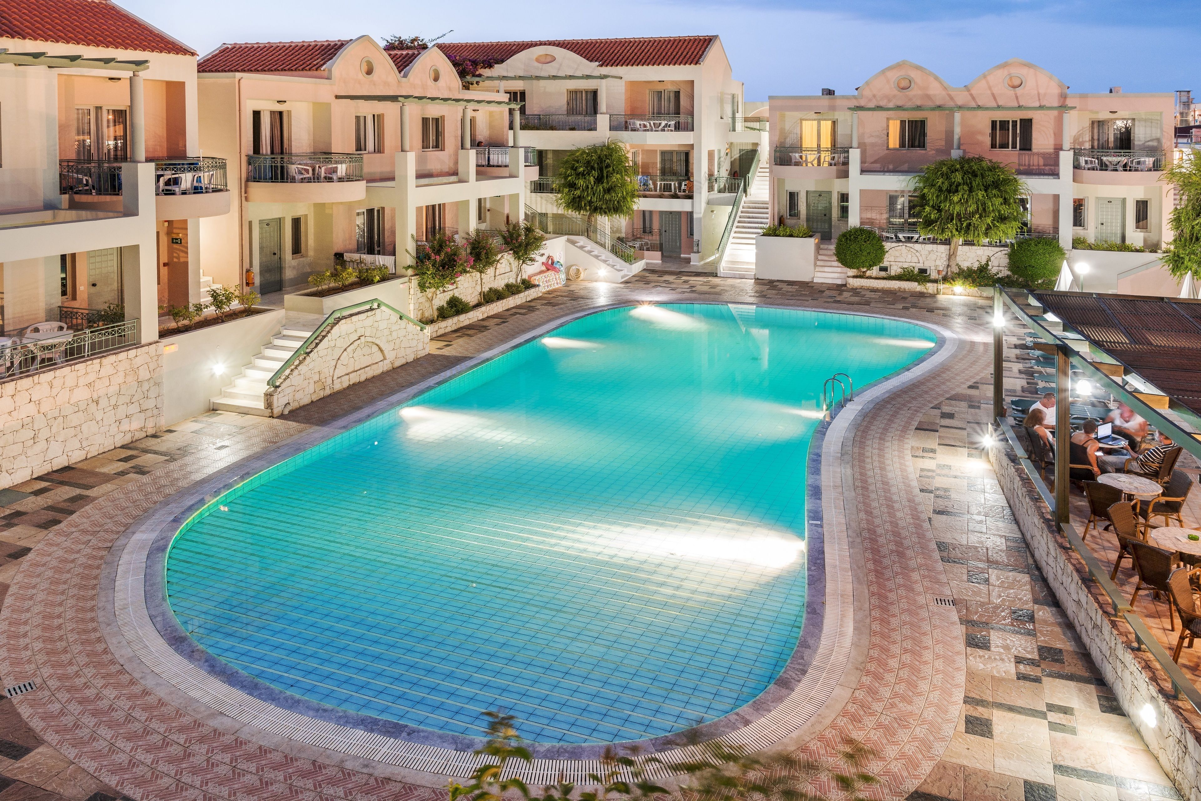 Ein Schwimmbecken, umgeben von mehrstöckigen Gebäuden mit Balkonen, einer überdachten Terrasse und einem Kiesweg, der zum Pool führt.