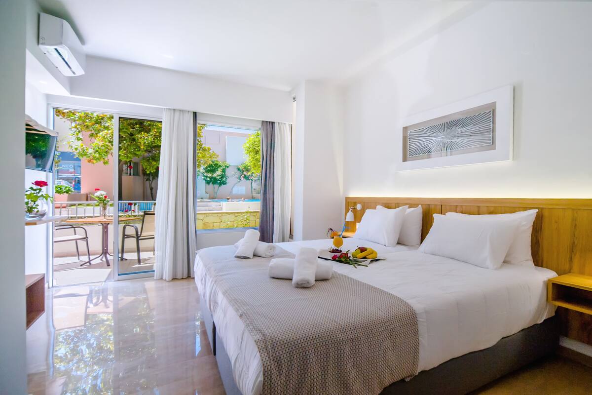 A hotel room with a large bed, a wooden headboard, a small table with a lamp, and a view of the outdoors through a sliding door.