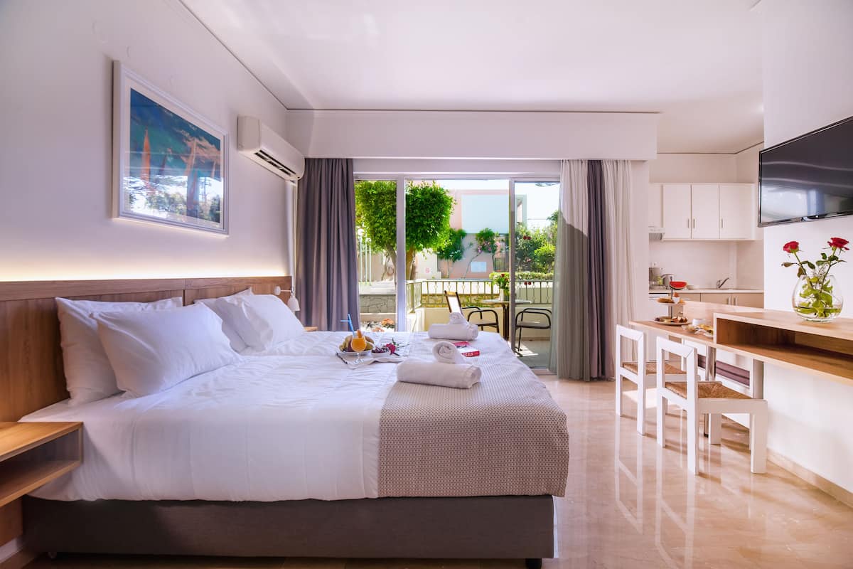 A hotel room with a large bed, a desk, a TV, and a balcony with a view of trees and buildings.
