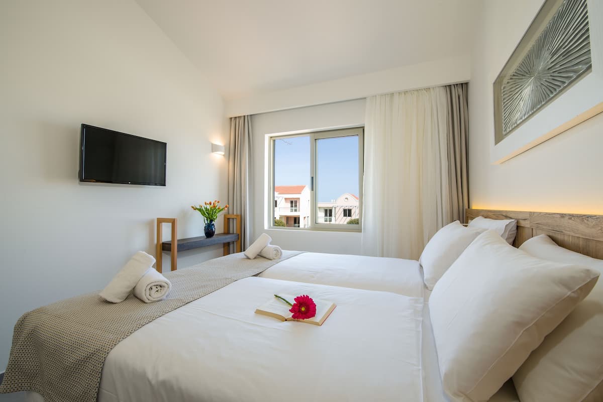 A hotel room with two beds, a TV mounted on the wall, a window with curtains, and a view of buildings outside.