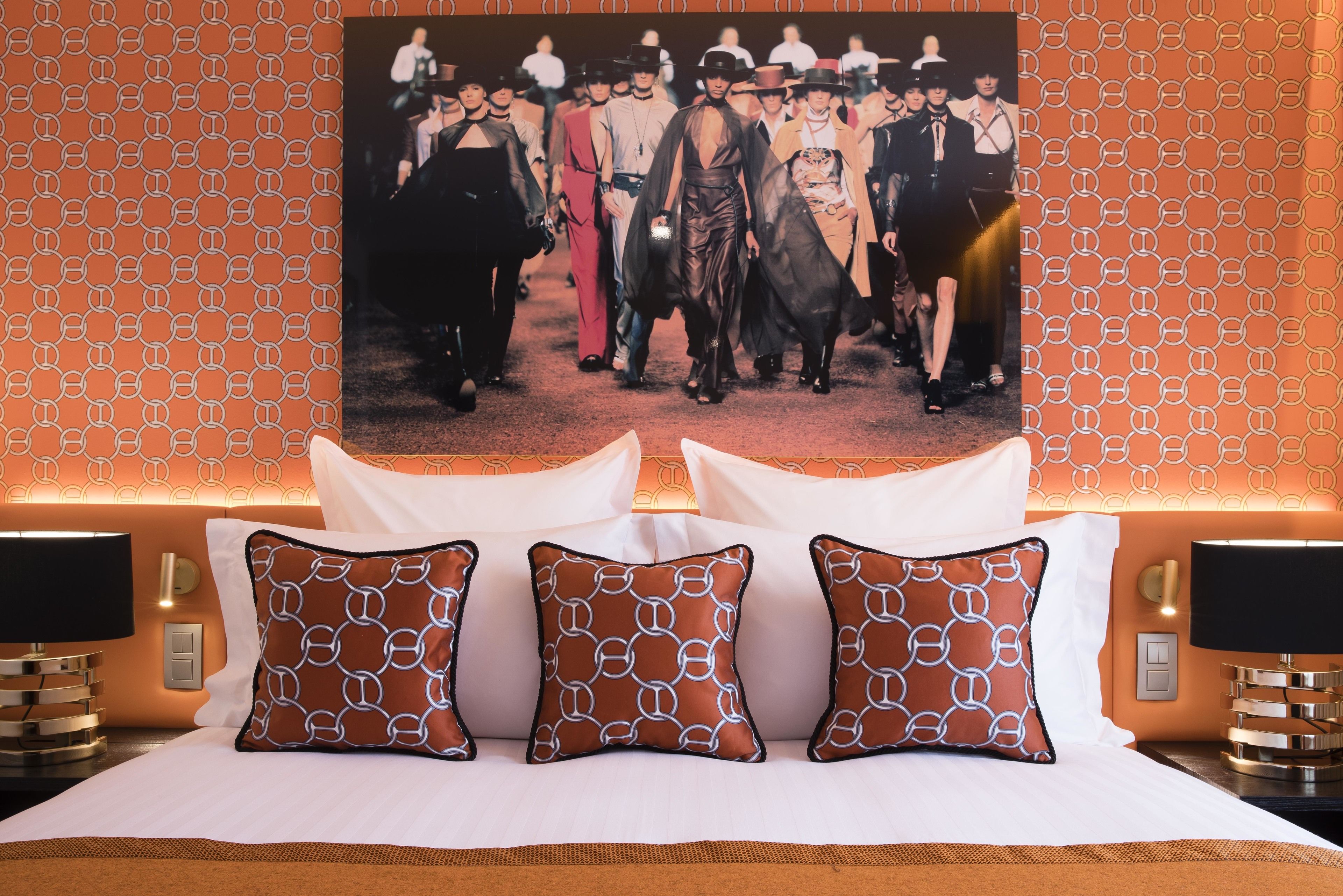 Quarto de hotel com papel de parede estampado, um pôster grande na parede e três almofadas estampadas na cama.