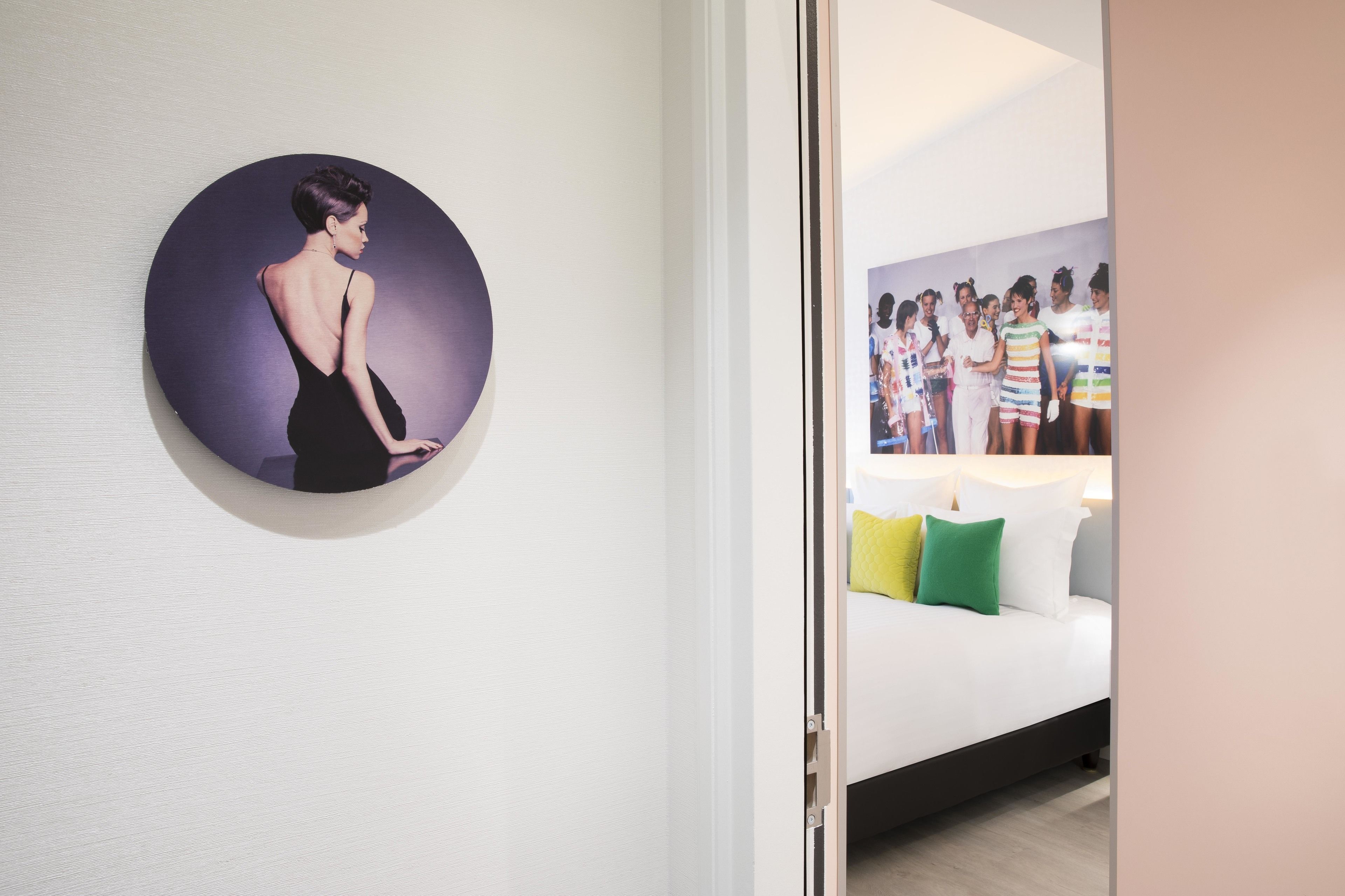 Un pasillo con una obra de arte circular en la pared, una puerta blanca y una cama con almohadas de colores al fondo.