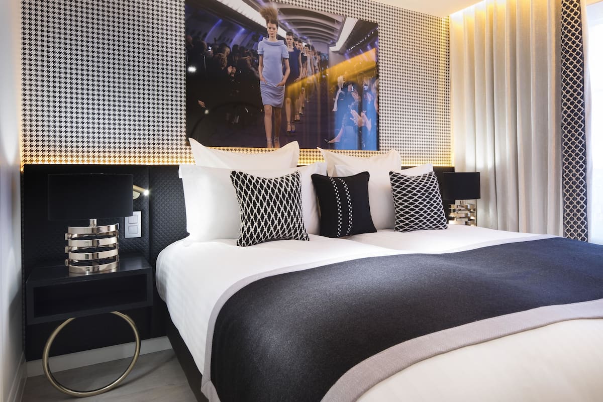 Habitació d'hotel moderna amb un llit gran, almoades amb dibuixos en blanc i negre, una nitzeta negra amb una làmpada i una televisió penjada a la paret que mostra un desfiladeu de moda.