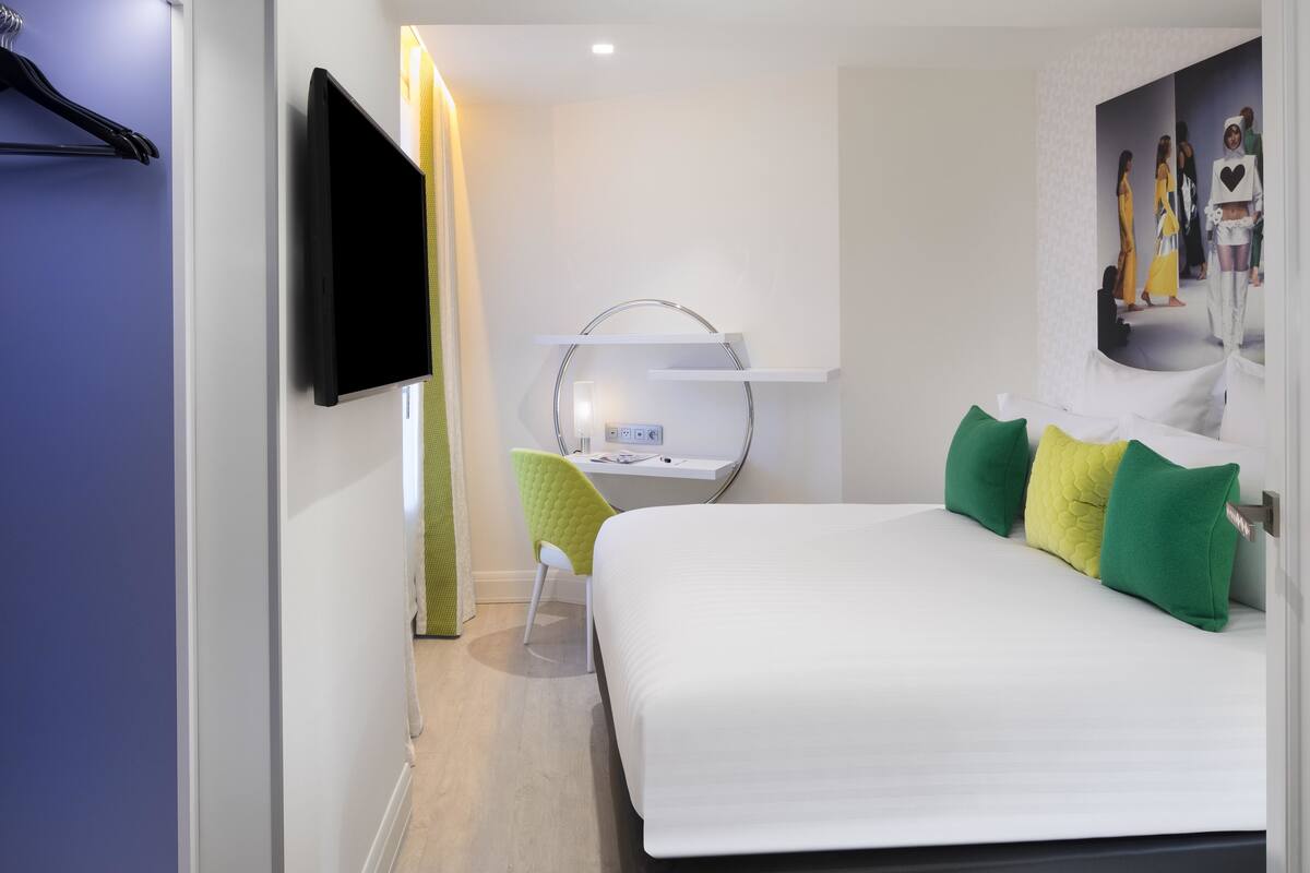 Habitació d'hotel moderna amb un llit, un escriptori, una cadira, un televisor i un mirall.