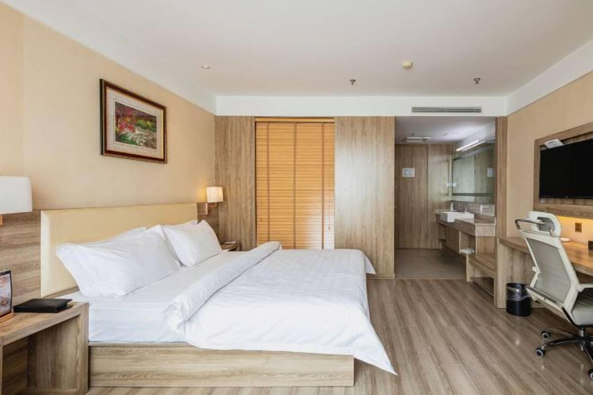 Une chambre d’hôtel avec un lit en bois, un bureau avec une chaise, une salle de bain avec un miroir et un tableau encadré au mur.