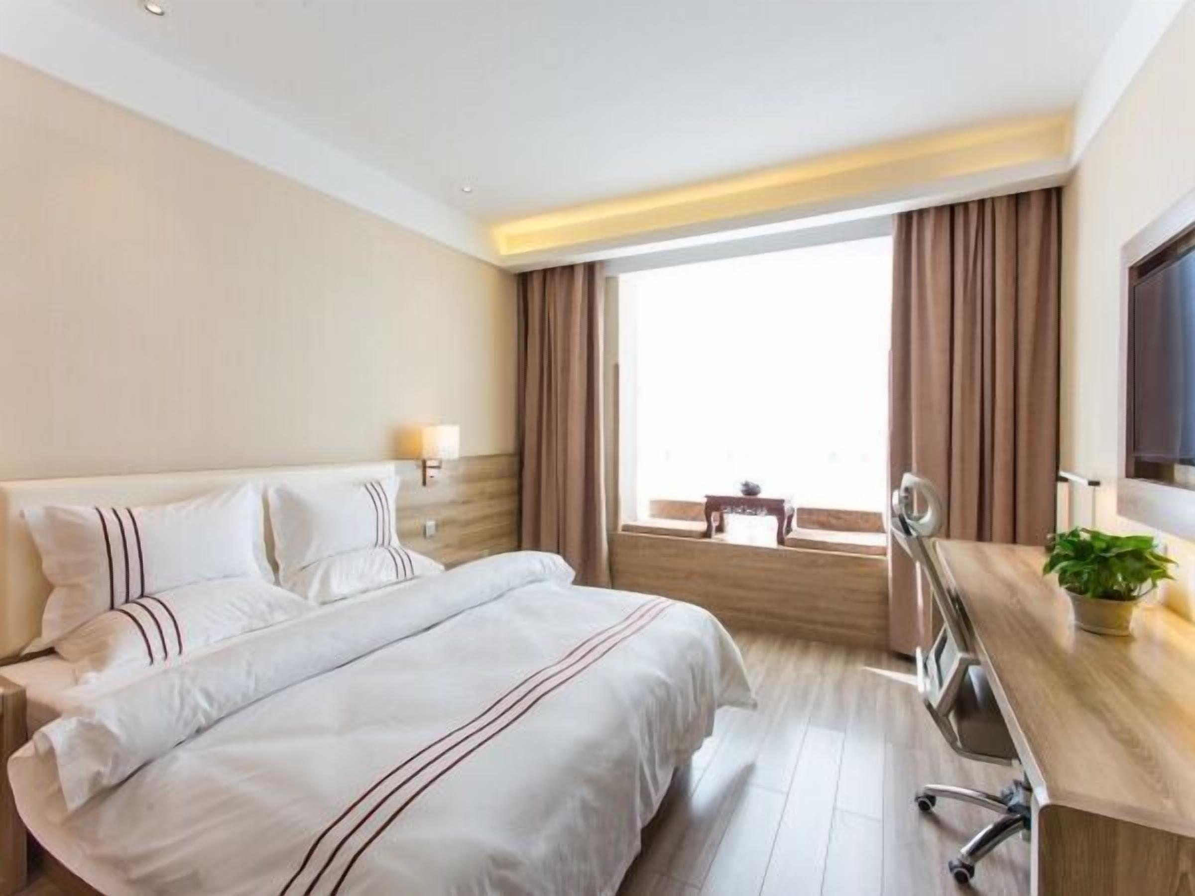 Habitación de hotel con una cama grande, un escritorio con silla, un televisor y un ventanal con cortinas.