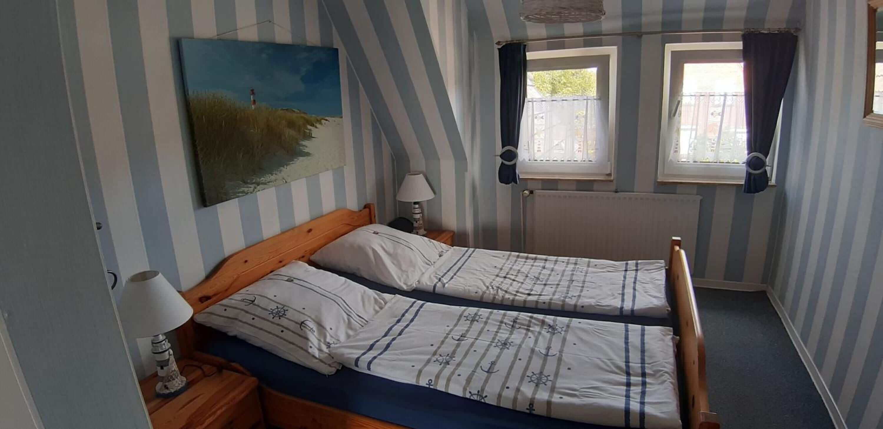 A room with two single beds, a nightstand with a lamp, a painting of a beach scene, and two windows with curtains.