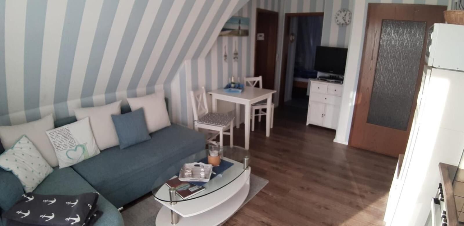 A compact living space with a sofa, a glass coffee table, a dining area with a table and chairs, a television, and a kitchenette.