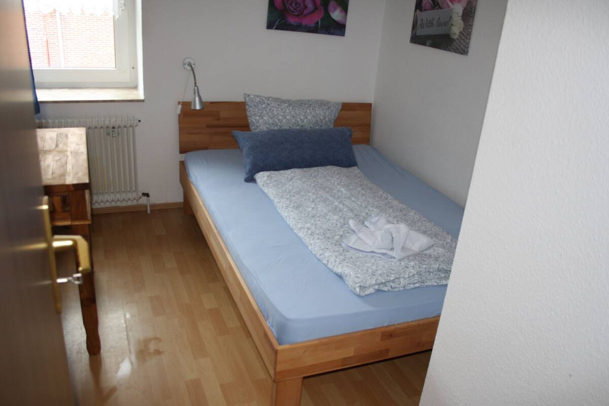 A single bed with a wooden frame, a blue mattress, and a white blanket. There is a wooden nightstand with a lamp, a radiator, and a window with blinds.