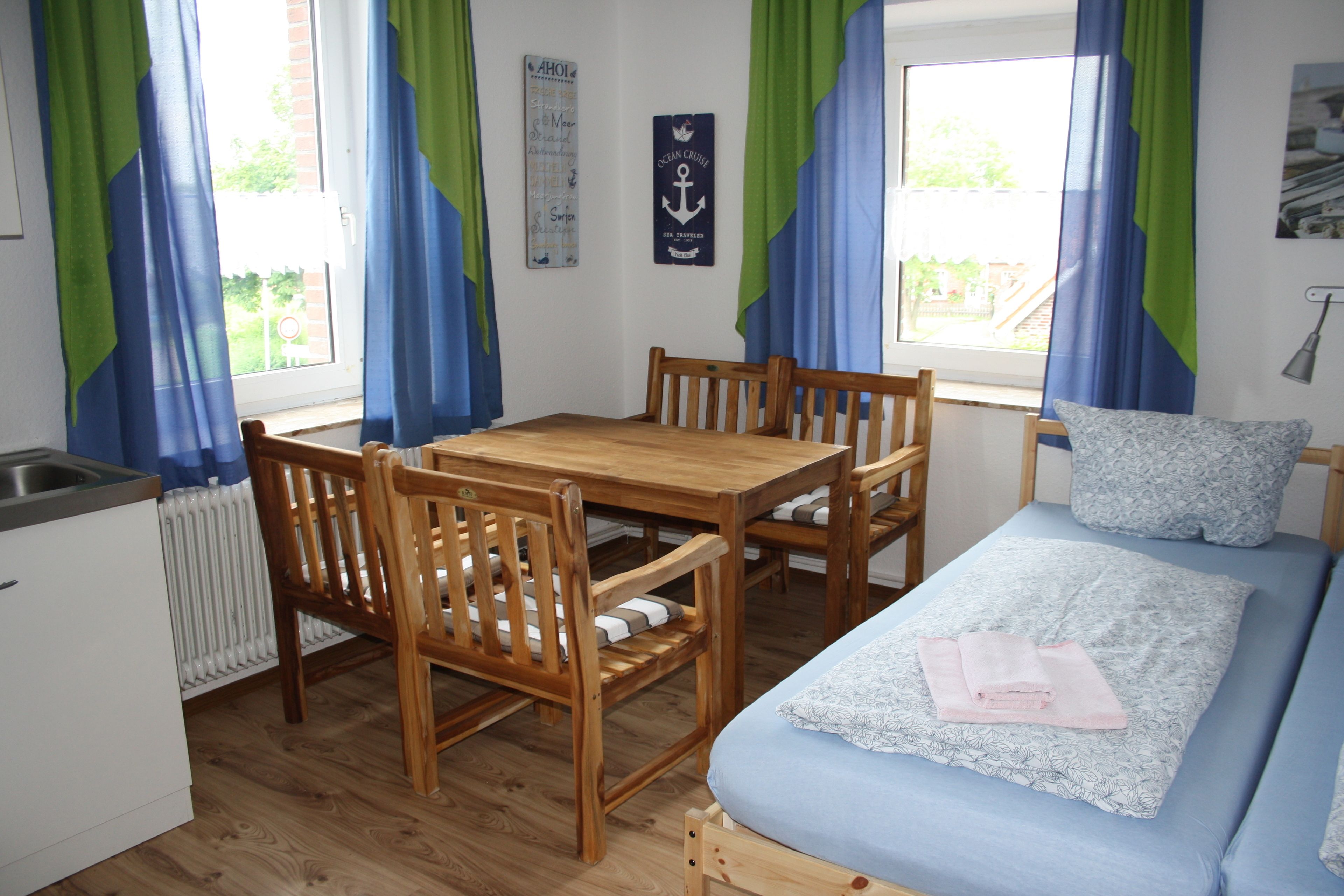 Una habitación compacta con una mesa de comedor de madera y sillas, una cama individual con colcha azul, y un ventanal con cortinas azules y verdes.
