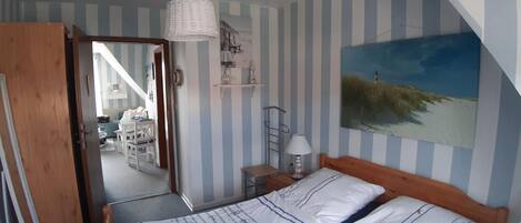 A bedroom with striped walls, a bed with a wooden headboard, a bedside table with a lamp, a painting of a beach scene, and a door leading to another room.