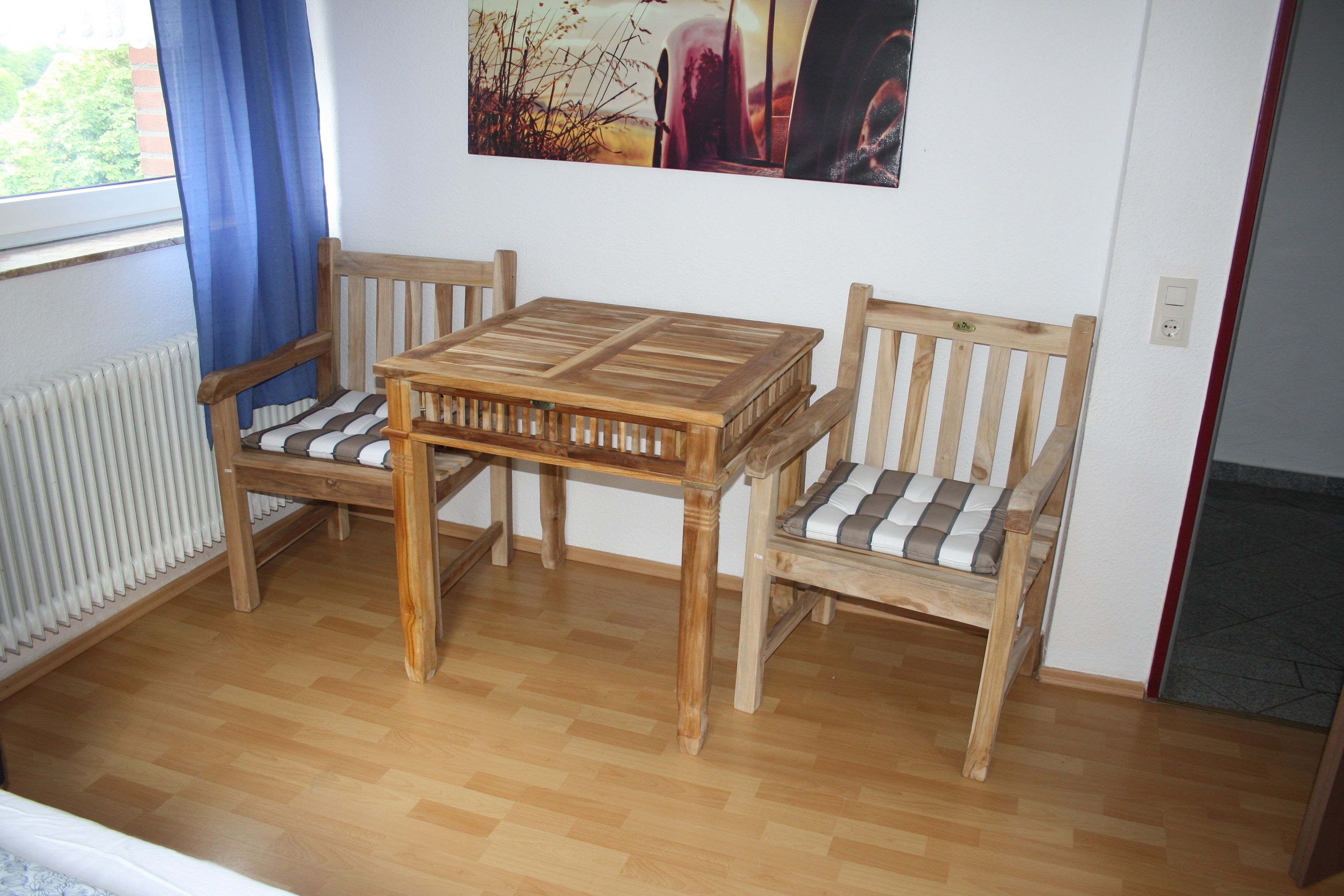 A room with a wooden table and two chairs, a radiator, a window with blue curtains, and a painting on the wall.
