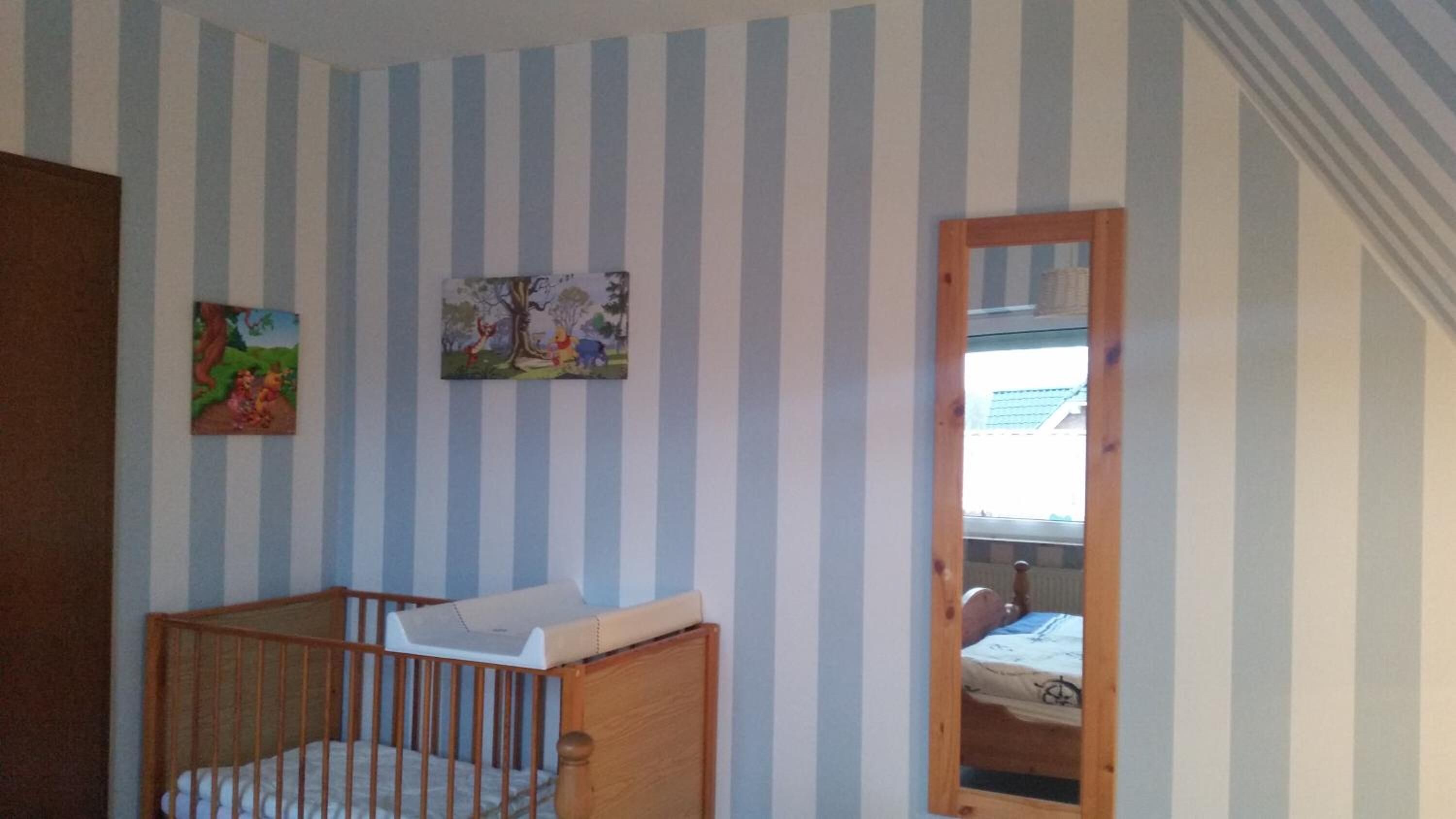 A room with striped walls, a crib, a bed, and a mirror reflecting a window.