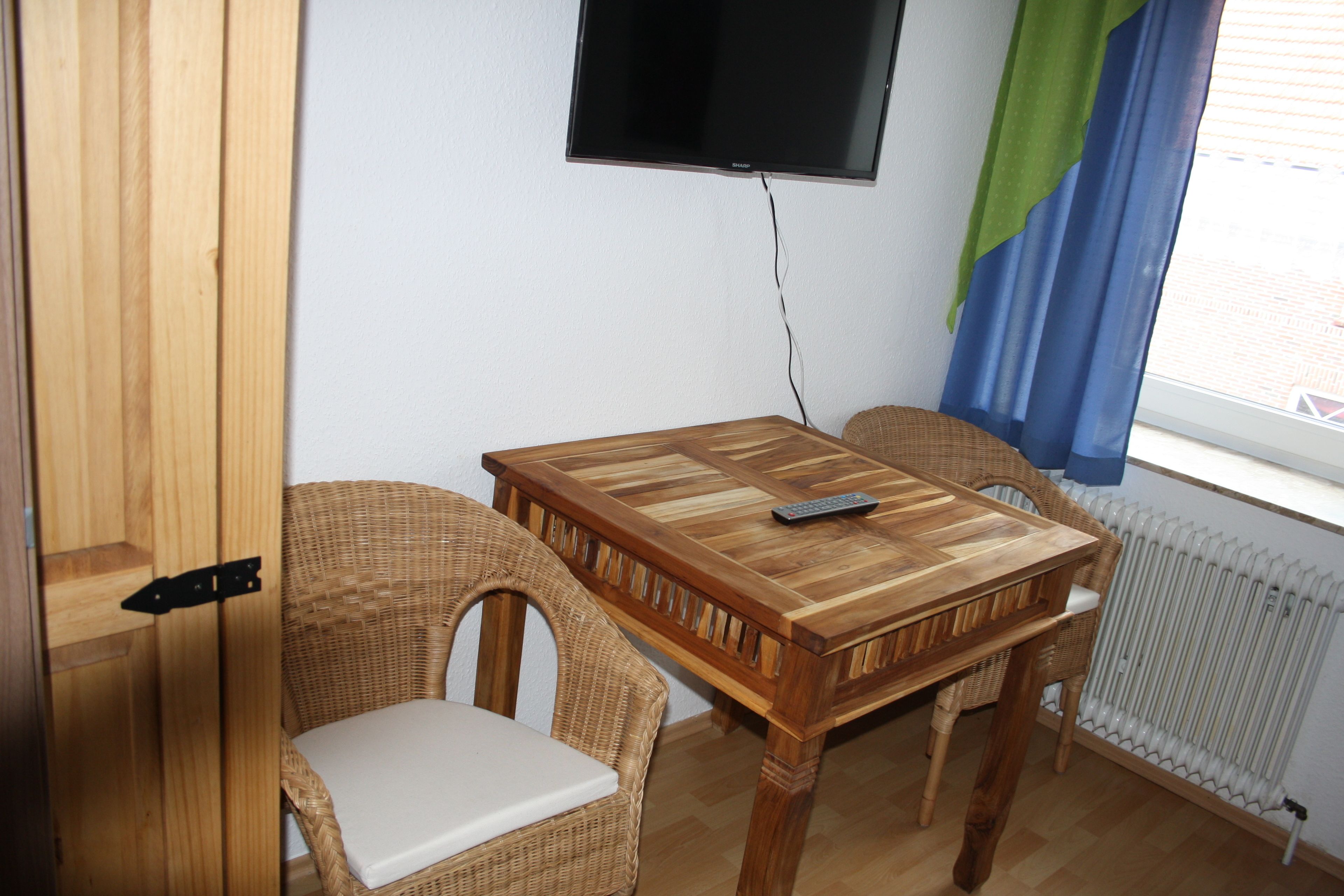 A room with a wooden table, two wicker chairs, a television mounted on the wall, and a window with a curtain.