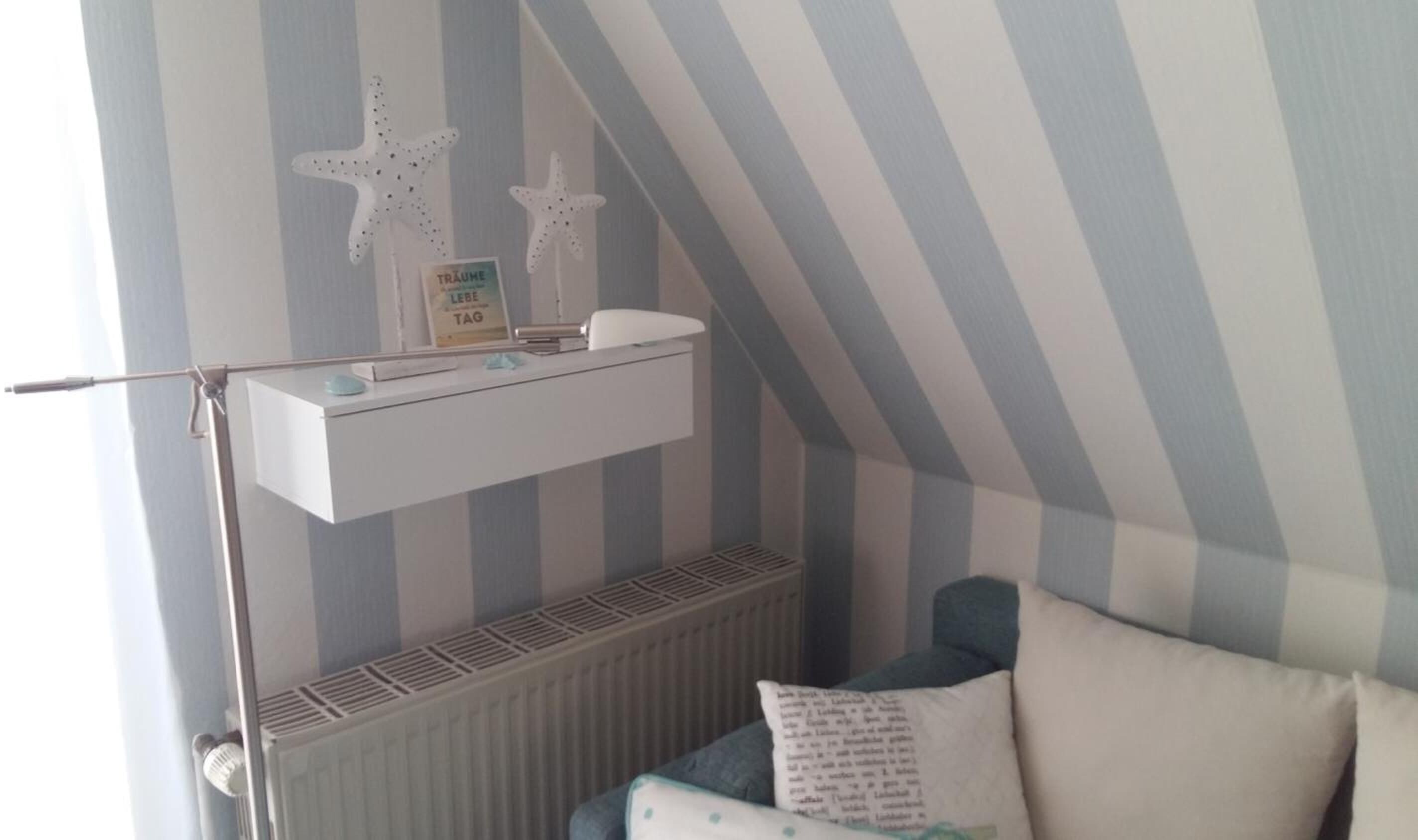 A room with a sloped ceiling, striped wallpaper, a white shelf with starfish decorations, a radiator, and a blue sofa with pillows.