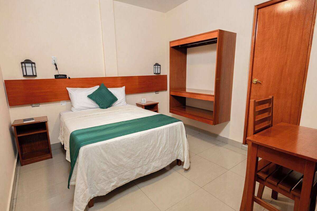 Una habitación de hotel con cama, mesitas de noche, un armario de madera y una pequeña mesa de madera.