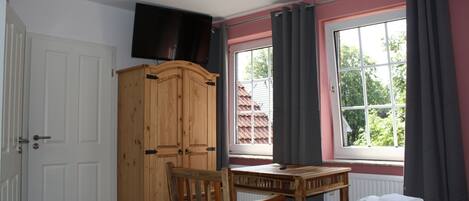 A bedroom with a wooden wardrobe, a desk, a chair, a bed with towels, a television on the wall, and a window with curtains.
