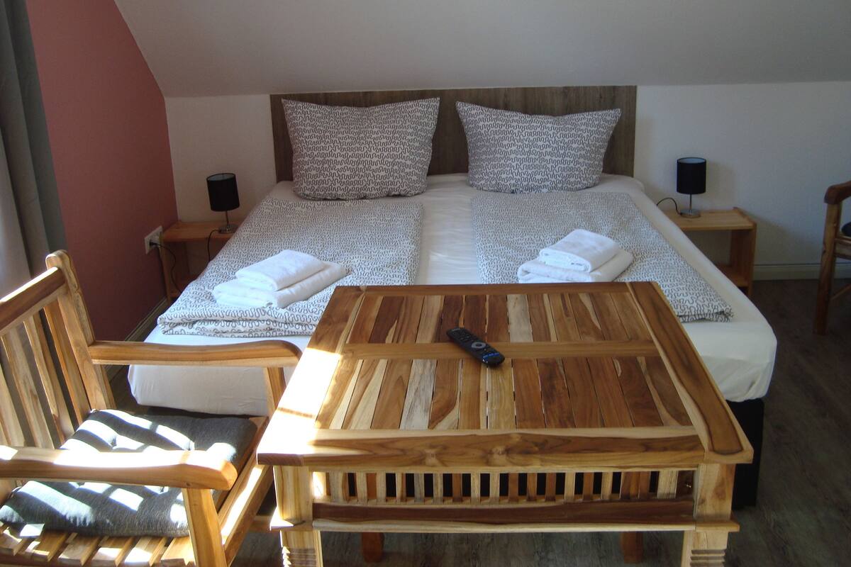 A bedroom with a bed, two pillows, a bedside table with a lamp, a wooden chair, and a table with a remote control.