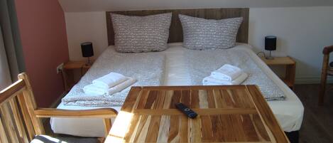 Un dormitorio con cama, dos almohadas, una mesita de noche con lámpara, una silla de madera y una mesa con control remoto.