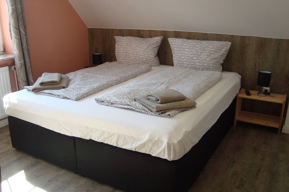 A double bed with a white duvet and patterned pillows, a wooden nightstand with a lamp, and a window with curtains.