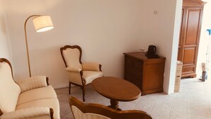 Una habitación con sofá, sillón y una pequeña mesa. Hay una lámpara de pie, un armario y un aparador de madera con una tetera y copas de vino encima.