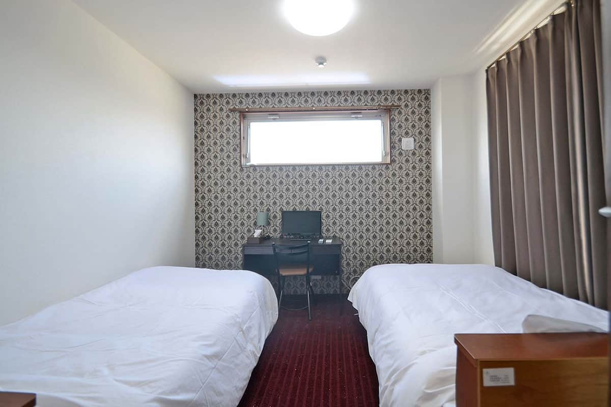A hotel room with two beds, a desk with a computer, a window, and a red carpet.