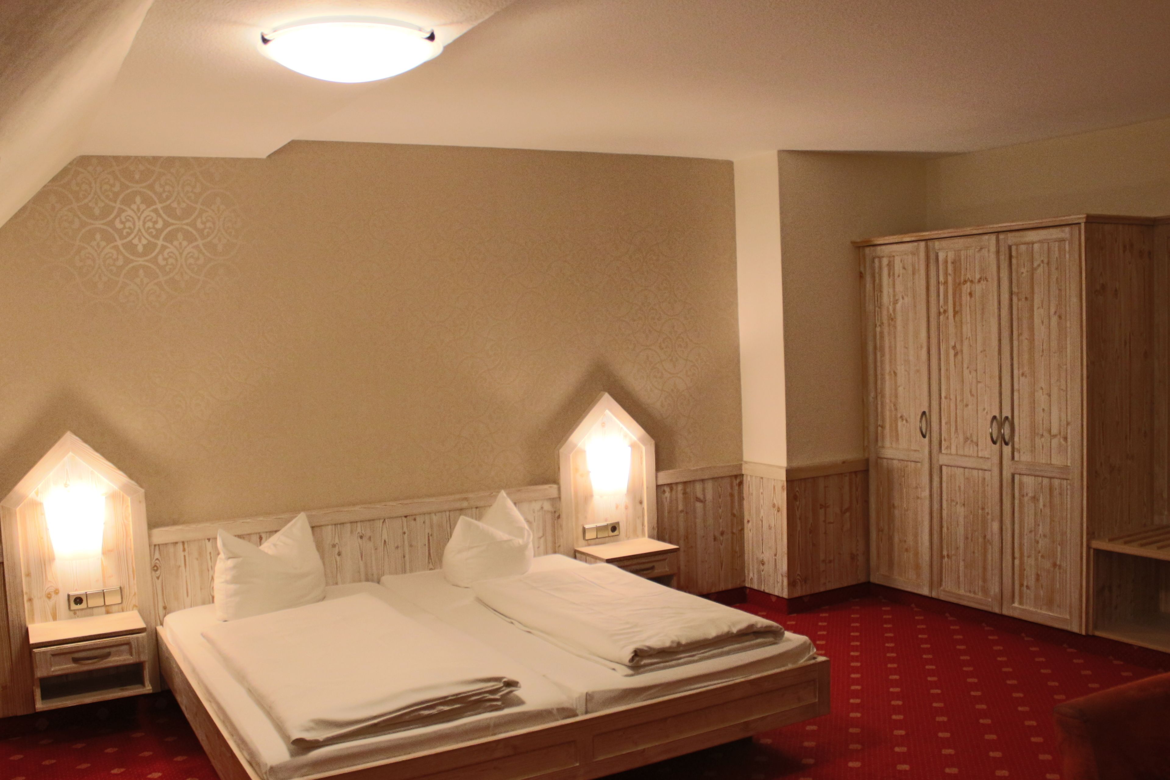 A bedroom with a large bed, two bedside tables, a wardrobe, and a sloped ceiling with a light fixture.