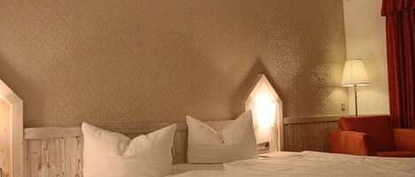 A neatly made bed with white linens and pillows, a wooden headboard, a nightstand with a lamp, and a red chair in a room with patterned wallpaper.
