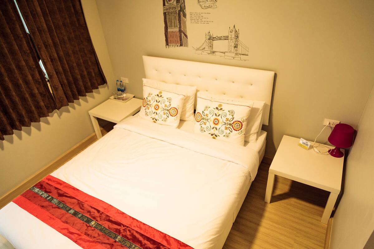 A neatly made bed with a red and white bedspread, two decorative pillows, a bedside table with a lamp, and a wall decoration of a bridge.