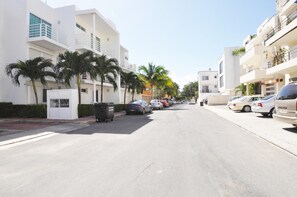 A street lined with modern buildings, parked cars, and palm trees.