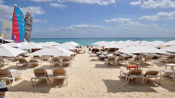 Une plage bordée de rangées de transats et de parasols blancs, un ciel bleu azur et une mer calme.