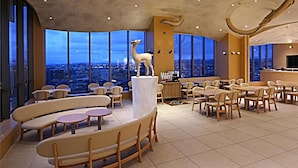 Un comedor moderno con techo alto, ventanas amplias que ofrecen vista a la ciudad y una estatua de jirafa como pieza central.