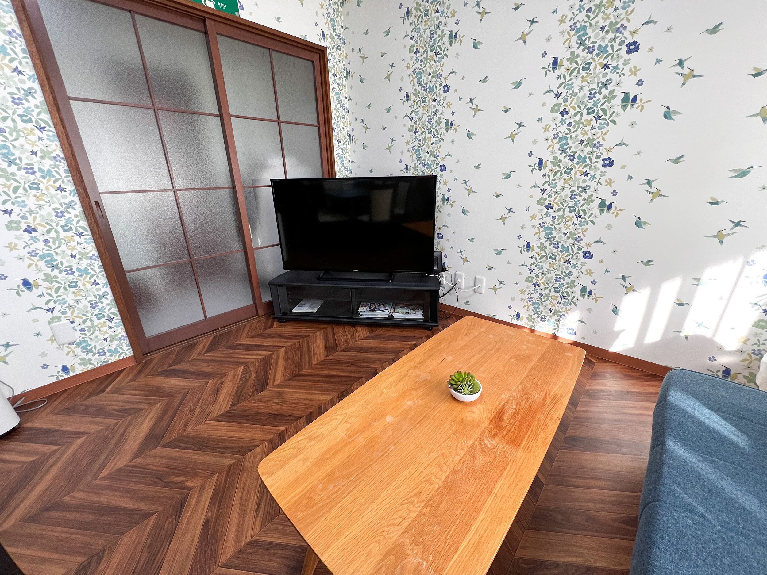 A room with a wooden herringbone floor, a television on a stand, a small plant on a wooden table, and floral wallpaper.