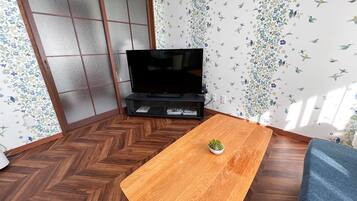 A room with a wooden herringbone floor, a television on a stand, a small plant on a wooden table, and floral wallpaper.
